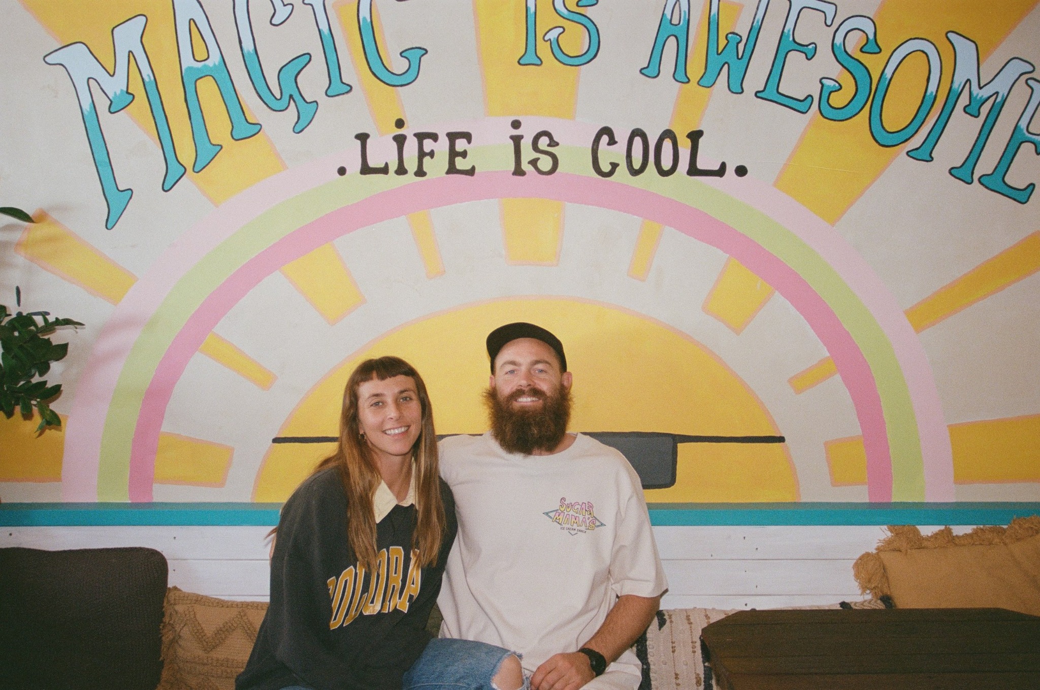 Kat and Timmy, founders of Sugar Mama's Shack, seated in front of the Magic is Awesome mural
